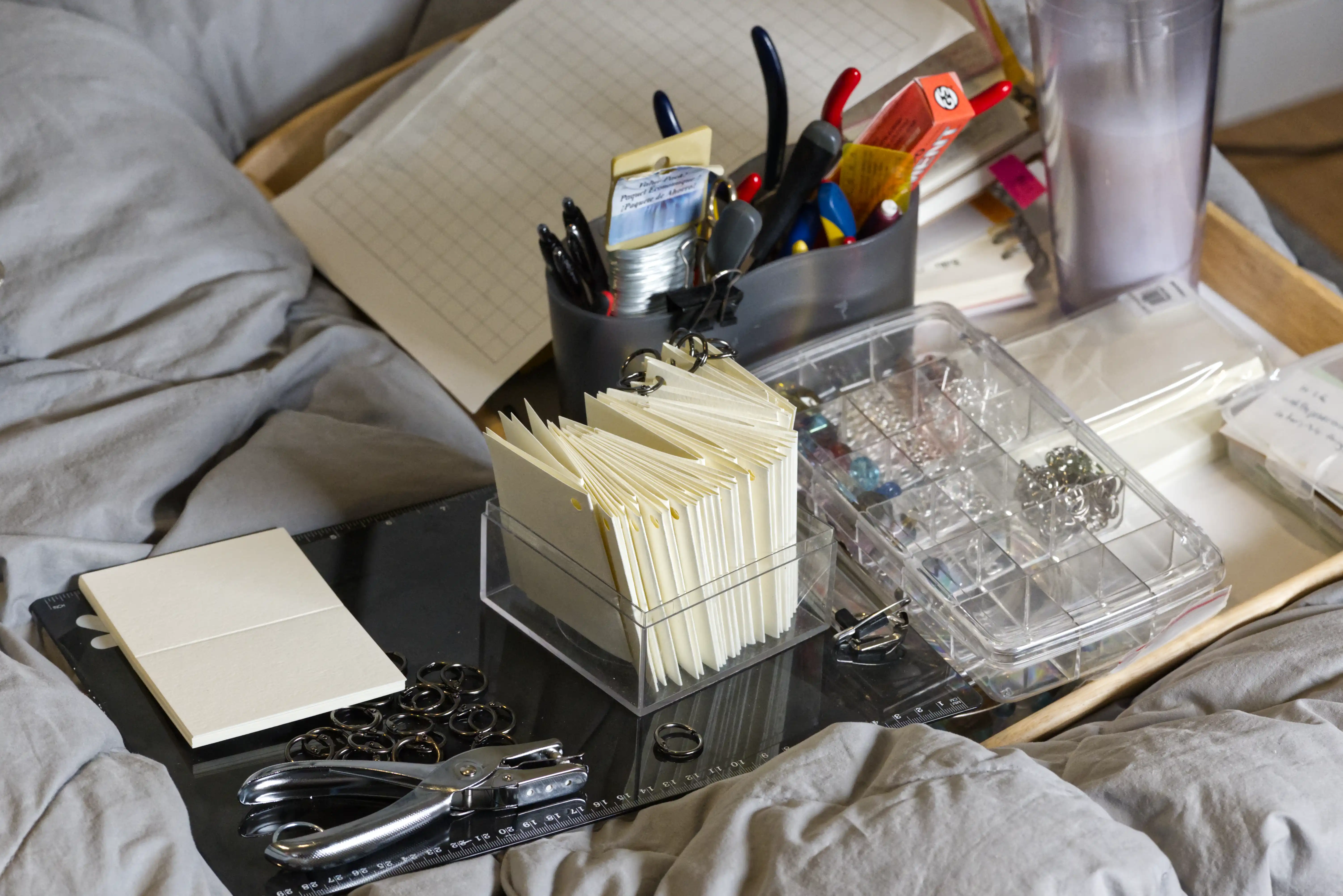 A surface that appears to be a bed, with crafting materials and tools atop: A hole punch, small booklets with punched holes, rings to hang on those holes, jewelerymaking tools, etc
